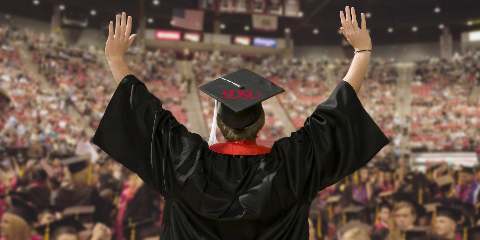 SDSU graduate in black cap and gown raising both arms in celebration